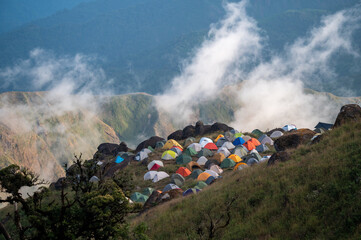A vibrant cluster of colorful tents set on a grassy mountain slope surrounded by mist, with...