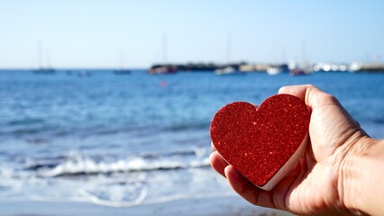 Fototapeta premium Happy Valentines day seaside. Hand holds red glitter heart in front of the sea. Sailboats on horizon in the blurred background. Love and romance concepts