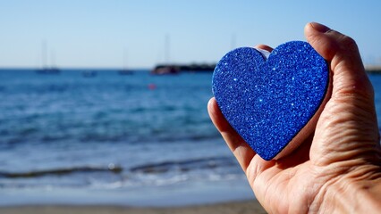Fototapeta premium Valentines day seaside. Hand holds heart with blue glitter in front of the sea. Sailboats on horizon in the blurred background. Love and romance concepts