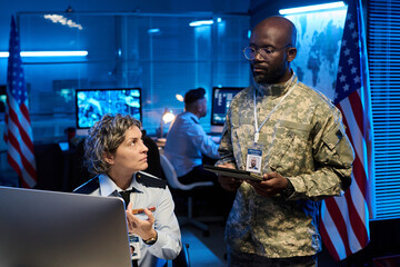Serious mature female officer in uniform pointing at computer screen and looking at young male colleague during discussion