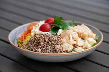 Close-up of a plate of raw vegetable salad standing on a table made of gray slats. The plate contains grains, cauliflower, carrots, tomatoes and cream cheese