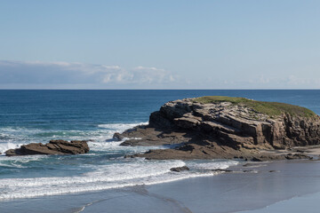 Coastal rock formation surrounded by ocean waves and a clear blue sky on a sunny day.