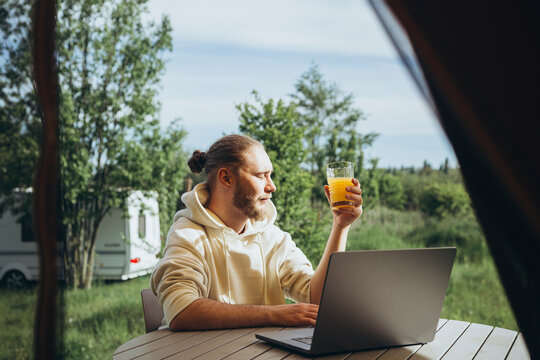 A young man drinks juice and works on a laptop in nature remotely.