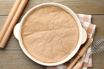 Ceramic casserole, baking parchment paper, brush and whisk on wooden table, flat lay