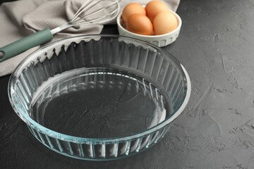Glass casserole, whisk and eggs on black table, closeup. Space for text