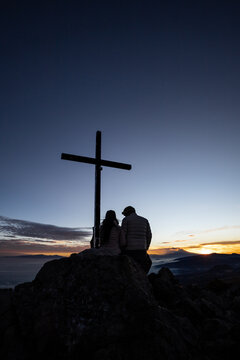 Couple watching sunrise next to cross on mountain peak