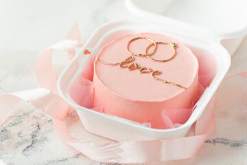 Little bento cake with pink cream cheese frosting decorated with golden wedding rings painting and love text. Beautiful anniversary cake on the white marble background