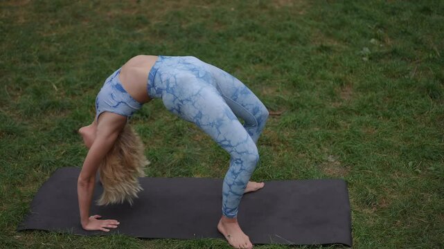 A blonde woman in a blue patterned yoga outfit performs a graceful backbend pose on a black mat in an outdoor setting. The scene captures her flexibility and strength amidst the natural greenery of
