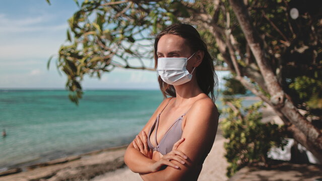 A woman in a face mask stands alone on a beach, the waves crashing behind her under the summer sun. Her presence reflects a stark contrast between serenity and the ongoing COVID-19 crisis.