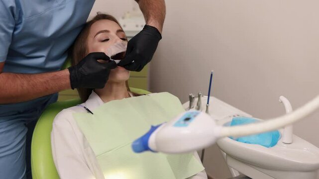 Dentist scrubs, Dental surgery, Dental treatment. Seated in dental chair, woman has her mouth held open by retractor while dentist in blue scrubs examines her teeth.