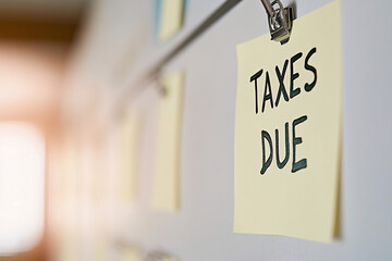 A minimalistic close-up of a yellow sticky note pinned on a board displaying the text taxes due in black, softly illuminated by a warm background light