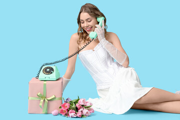 Young woman talking by retro telephone with gift box and bouquet of beautiful tulips on blue background. International Women's Day