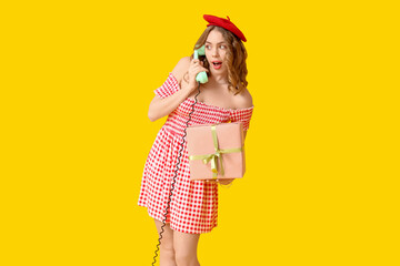 Young woman talking by retro telephone and gift box on yellow background. International Women's Day