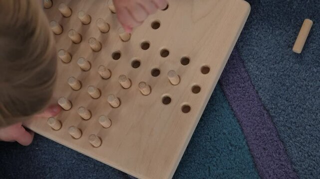 Young child plays with wooden solitaire game, top view. Toddler enhancing fine motor skills and concentration through engaging activities involving wooden pegs