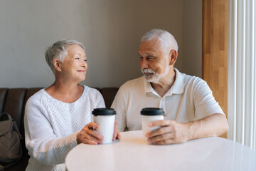 Happy elderly couple enjoying coffee and conversation at cozy cafe table sitting by window, exuding love and happiness, loving looking at each other, showing joy of togetherness in later years.