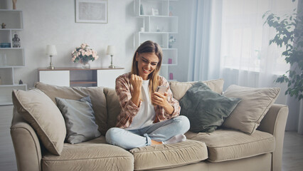 Overjoyed young woman is using mobile phone at home, sitting on comfortable sofa, reading good news in message, celebrating online win, achievement, receiving money prize, feeling excited and happy