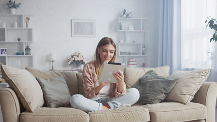 Smiling woman using digital tablet while sitting on comfortable sofa in cozy living room, enjoying online entertainment or working remotely