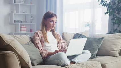 Obraz premium Comfortable casual millennial woman using laptop computer working remotely from home, sitting with legs crossed on sofa in cozy living room with natural light