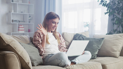 Happy young woman waving at her laptop during a video call while relaxing on a comfortable sofa in her living room, enjoying a moment of connection and communication from the comfort of her home