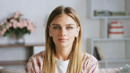 Confident young adult woman with blond hair and gold earrings sitting on a sofa in a cozy living room, looking directly at the camera with a neutral expression