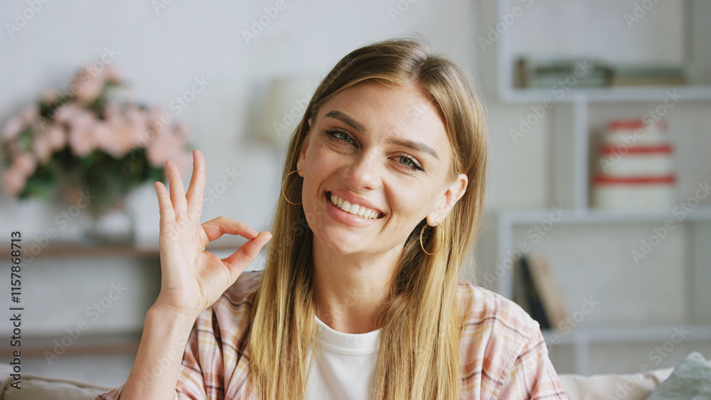 Wall mural Young, cheerful woman smiling and making an ok sign with her fingers while relaxing on a sofa in a cozy living room, enjoying the comfort of her home