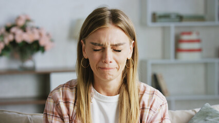 Young woman crying on the sofa at home, eyes closed and tears streaming down her cheeks, expressing...