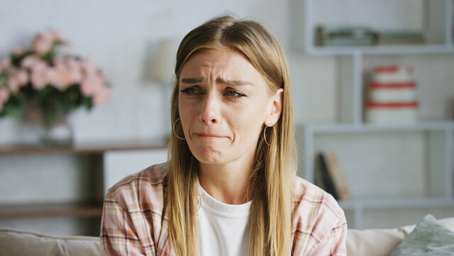Young woman sitting on a comfortable sofa at home is crying and wiping tears from her face, suffering from depression and mental health problems