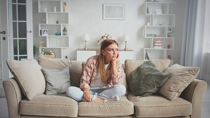 Sad woman sitting on the sofa at home, feeling lonely and overwhelmed, lost in thoughts about her...