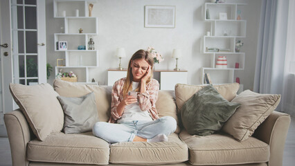 Obraz premium Young woman sitting on a comfortable sofa at home, reading bad news or receiving upsetting message on smartphone and feeling worried, stressed, frustrated, or tired