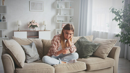 Young woman sitting on comfortable sofa at home reading bad news on mobile phone, feeling worried and stressed, having problem with online communication or notification