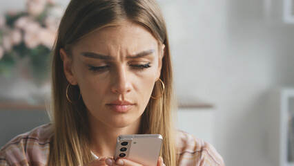 Young woman frowning while reading upsetting message on smartphone, experiencing disappointment and frustration with bad news, feeling stressed and anxious about negative online notification