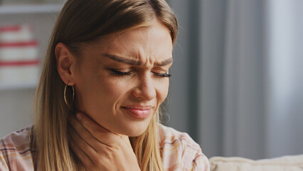 Close up of a young blond woman with closed eyes touching her neck, suffering from sore throat or...