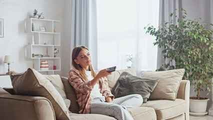 Bored woman sitting on a comfortable sofa in her living room, holding a remote control and changing...