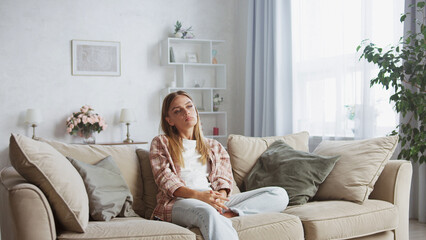 Young woman sitting on a cozy sofa in her living room, feeling bored and unmotivated while enjoying a lazy weekend day at home