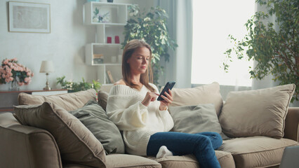 Young woman sitting comfortably on a cozy sofa in her living room, browsing her smartphone and enjoying a peaceful moment at home while relaxing in casual attire