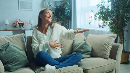 Obraz premium Cheerful woman is sitting on a comfortable sofa in her living room, gesturing with her hands and enjoying her weekend, feeling relaxed and happy in the cozy atmosphere of her home