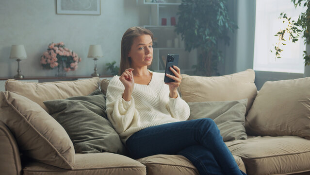 Young woman sitting comfortably on a cozy sofa in a serene living room, using her smartphone to enjoy a relaxing moment filled with connectivity and digital engagement