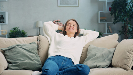 Smiling woman is lying on comfortable sofa with her hands behind her head, enjoying her leisure time in cozy living room, feeling relaxed and happy