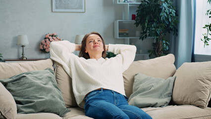 Fototapeta premium Happy woman enjoying her leisure time, lying on a comfortable sofa with her hands behind her head, eyes closed, and a peaceful expression on her face, surrounded by the cozy ambiance of her home