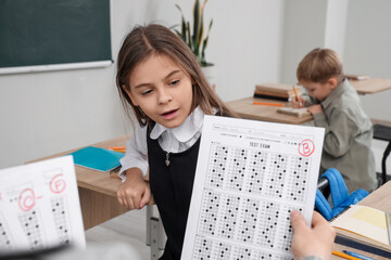 Cute schoolgirl receiving answer sheet with B grade from teacher in classroom
