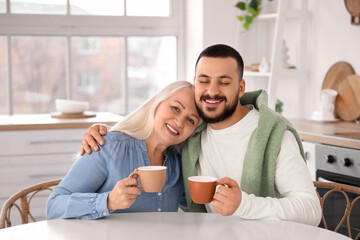 Young man and his mother with cups of tea hugging at table in kitchen