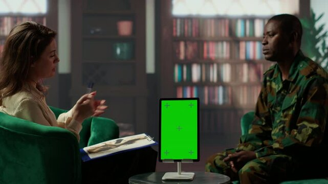 Infantry serviceman attending a therapy session with psychologist next to mockup display, suffering from PTSD after combat. Female counselor offers help and support to army officer. Camera B.