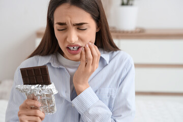 Young woman with chocolate bar suffering from toothache in bedroom, closeup