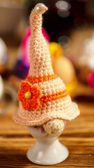 A creatively designed crochet gnome hat with distinct orange stripes and yellow flower embellishment, sitting playfully atop a small white ceramic holder, exuding charm.