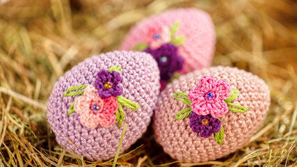 A collection of three pastel-colored crocheted eggs, each decorated with delicate floral embroidery, laying on natural hay, portrays a springtime festive theme.