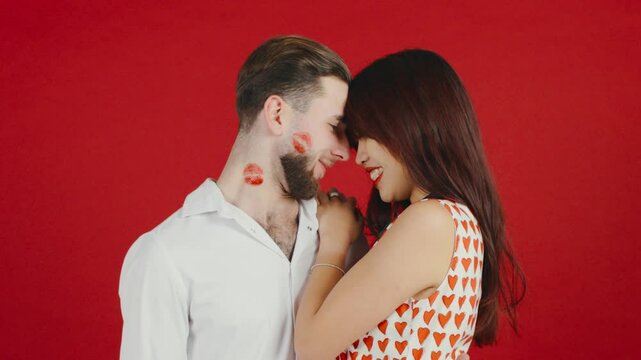 Man with kiss marks with red lipstick on his cheek and neck hugs his Asian girlfriend on a red background celebrating Valentines Day
