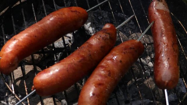 Sausages sizzle on a suspended metal grid above a circular tray, surrounded by a lively, possibly evening street food scene with rich aromas and flavors