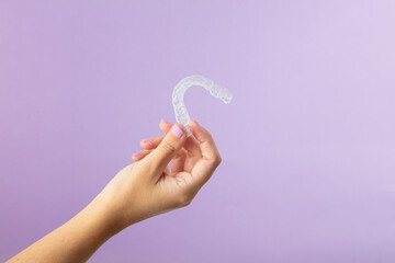 Close-up of a hand holding a transparent dental aligner against a soft purple background. The image highlights modern orthodontic technology and dental care solutions.