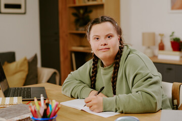 Medium portrait of confident young woman with disability sitting at wooden desk in living room, looking at camera while writing in notebook