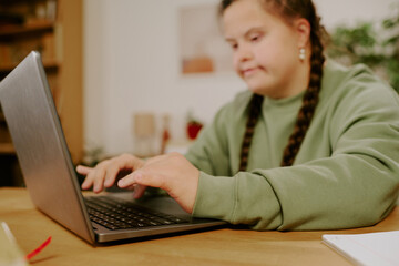 Medium closeup of young Caucasian girl with Down syndrome sitting at wooden table at home, typing on laptop